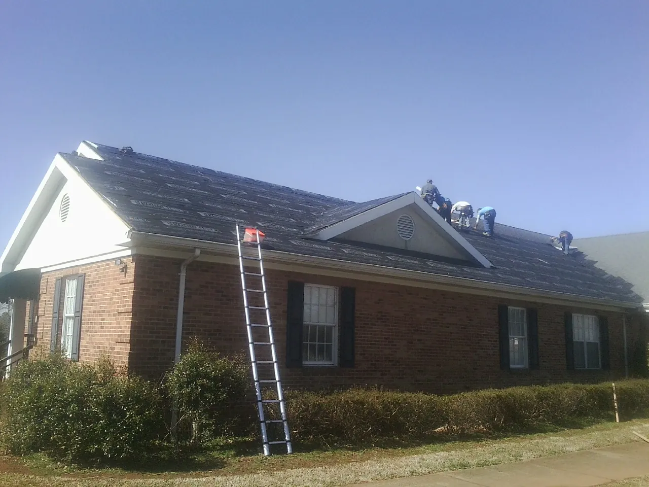 Crew tearing shingles off a brick ranch roof with a ladder set — Metal Roof Repair work by our crew in Kiel
