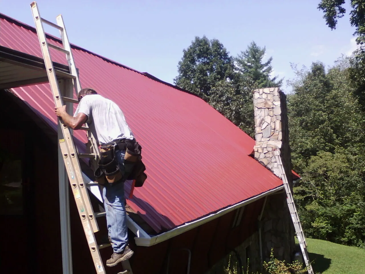 Roofer on a ladder beside a red metal roof and stone chimney — Soffit & Fascia Repair by Roofing Brothers in Kiel